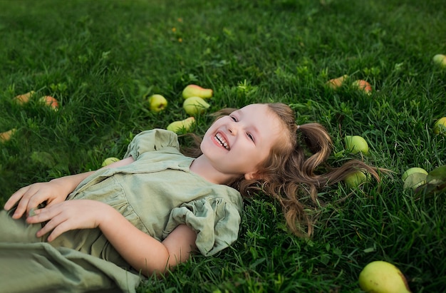 belle petite fille heureuse dans une robe verte est allongée sur l'herbe avec des fruits et rit