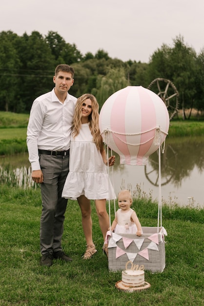 Une belle petite fille avec maman et papa dans un panier avec un ballon et un gâteau fête son premier anniversaire