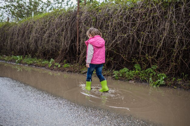 Belle petite fille marchant dans la rue et en bottes de caoutchouc vert sur des flaques d'eau tirant par derrière. La jeune fille marche en bottes et en pied bot. Le problème avec les pieds, la maladie des pieds.