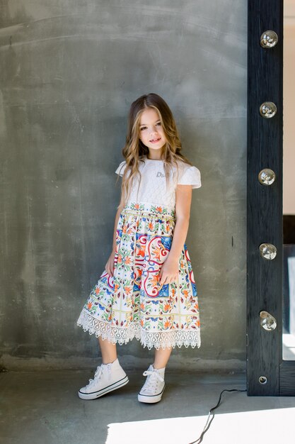 Belle petite fille mignonne dans un chemisier brillant rose et une jupe bleue sur un mur gris, debout près du miroir.