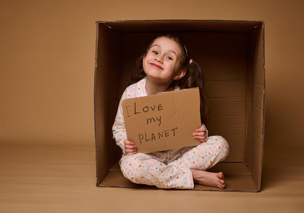 Belle petite fille militante tenant une pancarte avec Love my Planet assise à l'intérieur d'une boîte en carton en posture de lotus et souriante en regardant la caméra sur fond beige avec espace de copie