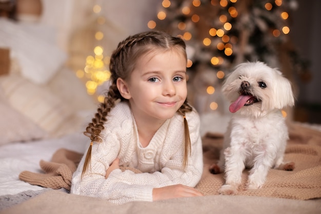 Belle petite fille a avec des nattes sur la tête joue avec son chien maltais sur le lit à Noël