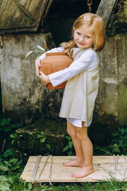 belle petite fille avec un pot et un seau près du puits sur le fond des arbres et des herbes