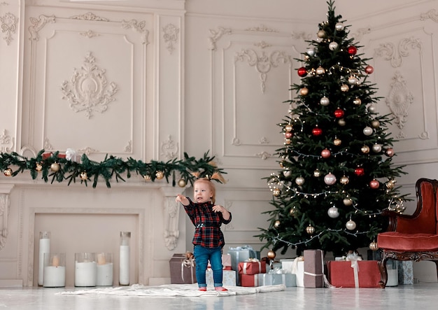 Belle petite fille avec des queues de cheval se tient près d'un arbre de Noël avec des cadeaux et des spectacles sur le côté