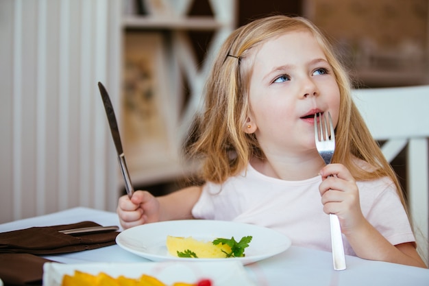 Belle petite fille en riant la blonde assise à une table dans un café et a mangé de la purée de pommes de terre dans l'assiette.