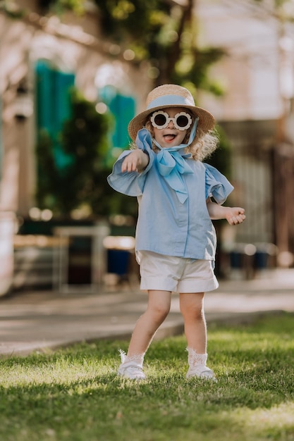 belle petite fille en robe bleue, chapeau de paille et lunettes de soleil jouant en plein air, fond bleu, carte