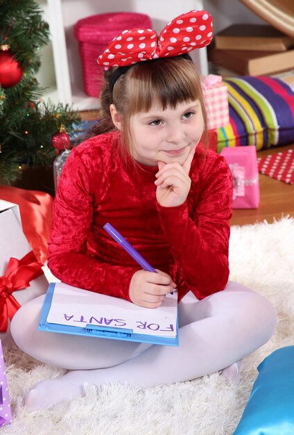 Belle petite fille en robe rouge écrit une lettre au Père Noël dans une pièce décorée de façon festive