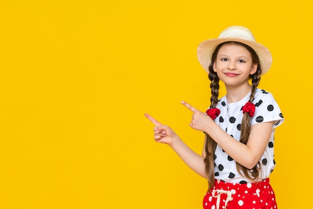 Une belle petite fille en short à pois et un chapeau de paille pointe vers votre publicité Vacances d'été Une adolescente attend l'été sur un fond jaune isolé Espace de copie