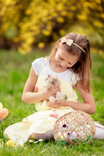 Belle petite fille souriante tenant un lapin mignon et assise sur l'herbe pendant les vacances de printemps.