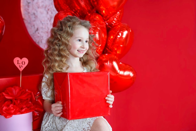 Belle petite fille sourit sur un fond rouge avec un ballon en forme de coeur et une boîte-cadeau dans ses mains Le concept de cadeaux d'amour Saint Valentin 14 février 8 mars fête des mères