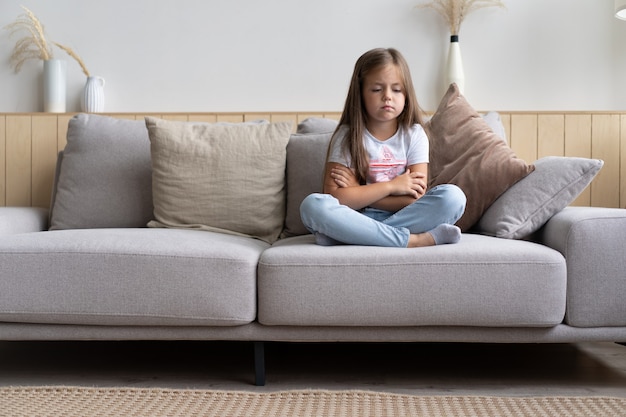 Belle petite fille triste et malheureuse de 6 ans assise les jambes croisées sur un canapé dans le salon, se sentant ennuyée et seule, étant seule à la maison pendant que ses parents passent toute la journée au travail
