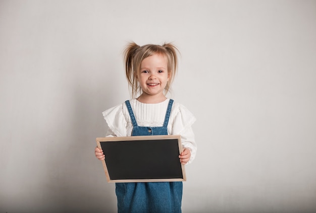 Une belle petite fille vêtue d'un chemisier blanc et d'une salopette en jean et d'une planche en graphite