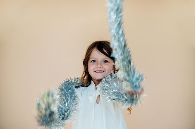 Une belle petite fille vêtue d'une robe blanche avec des guirlandes festives dans ses mains Le concept de vacances Joyeux Noël Nouvel An Les cheveux longs flottent la fille sourit et se réjouit