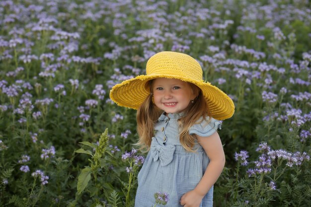 Une Belle Petite Fille Vêtue D'une Robe Bleue Et D'un Chapeau Jaune Se Tient Dans Un Champ De Fleurs