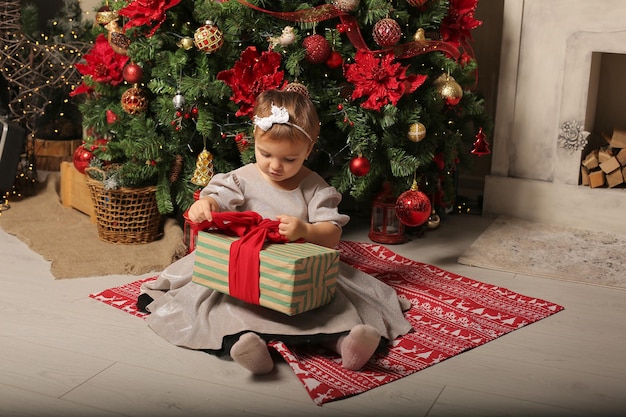 belle petite fille vêtue d'une robe de fête est assise près de l'arbre de noël à côté d'une boîte-cadeau