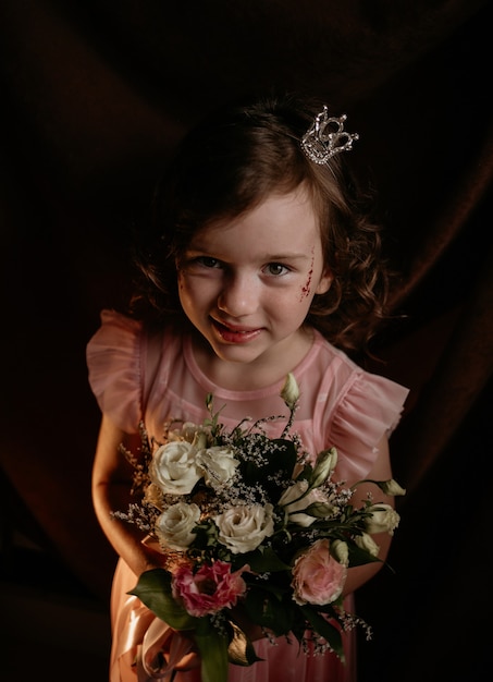 Belle petite princesse aux cheveux bouclés avec une couronne sur la tête tient un bouquet de fleurs sur un fond de tissu marron