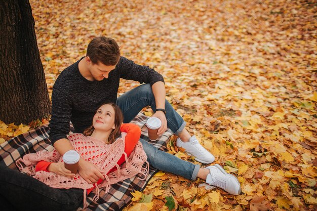 Belle photo de jeune couple dans le parc. Femme allongée sur une couverture et regardez le gars. L'homme s'assoit et regarde la femme. Ils ont des tasses de café.