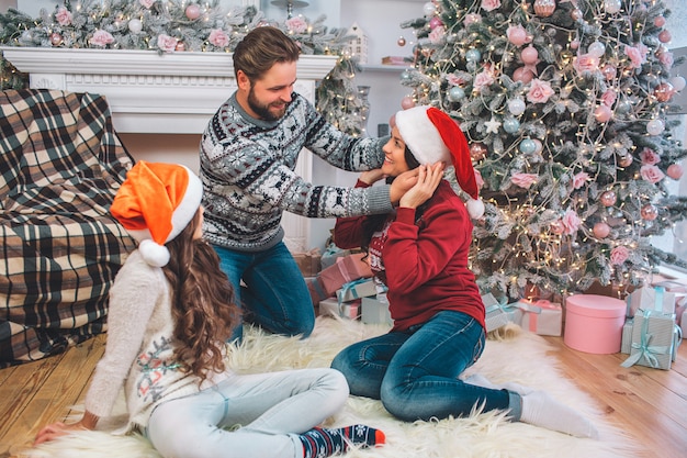 Belle photo d'un jeune homme debout sur les genoux devant la femme et mettant un chapeau rouge sur sa tête. Ils se sourient. Leur enfant est assis à côté d'eux. La famille a l'air festive.