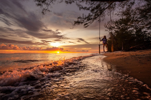 Belle plage tropicale au coucher du soleil, la fille est le bonheur se balançant sur une balançoire.