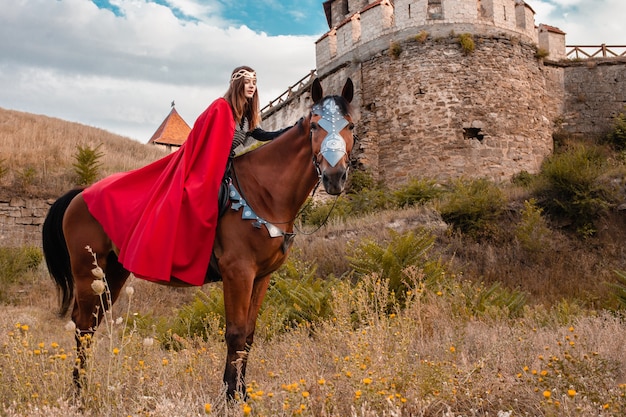 Belle princesse avec cape rouge à cheval dans le contexte d'une tour et d'un mur de pierre