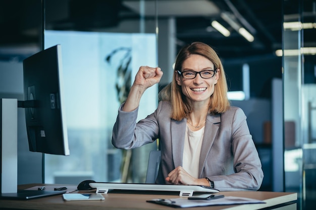 Belle et réussie femme d'affaires avec des lunettes célèbre la victoire regarde la caméra et se réjouit d'un projet achevé avec succès garde ses mains levées du bonheur et de la satisfaction