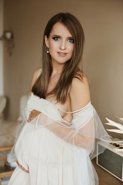 Belle et sensuelle brune jeune femme enceinte aux yeux bleus et avec un corps sexy en élégante robe beige posant à l'intérieur