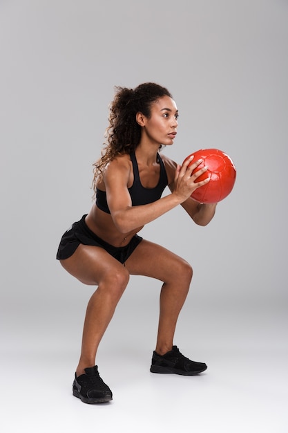 Belle sportive africaine isolée sur fond gris, faire des exercices avec un ballon lourd, accroupi