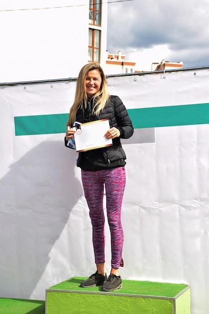 Une belle sportive blonde de la marche nordique sur un piédestal montre le prix gagné et sourit