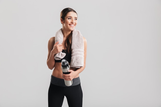 Belle sportive en bonne santé portant un survêtement avec une serviette en regardant de côté et tenant une bouteille d'eau fraîche, isolée sur un mur gris
