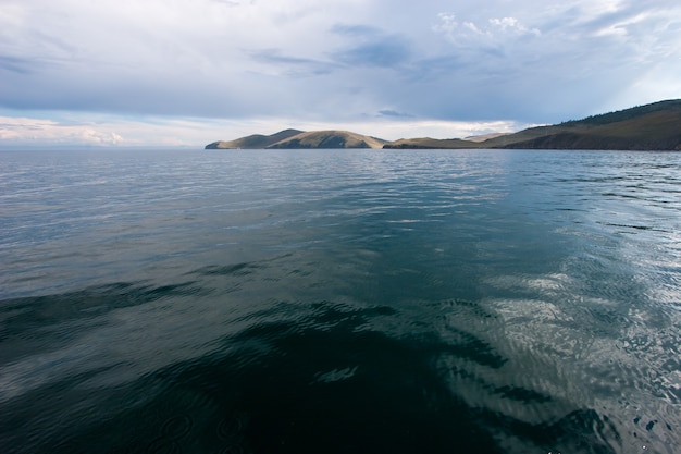 Belle vue sur l'eau du lac Baïkal par jour avec des nuages dans le ciel. Vue depuis l'eau. L'eau est verte. A l'horizon, des collines.