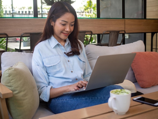 De belles femmes asiatiques utilisent des ordinateurs pour se connecter à Internet afin de communiquer avec des amis étrangers Une femme heureuse et détendue est assise sur le canapé Femme qui travaille et concept indépendant