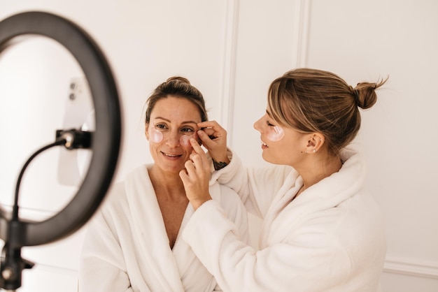 De belles femmes caucasiennes en peignoirs appliquent des patchs transparents d'hydrogel sous les yeux sur fond blanc. Journée avec soins de la peau et santé.