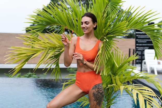 Belles femmes européennes en maillot de bain bikini orange deux pièces en parfait état, bon corps bronzé avec des lunettes de soleil à la mode à l'extérieur à la piscine tropicale et au palmier