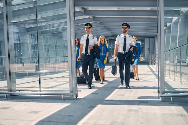 Belles hôtesses de l'air et beaux pilotes portant des sacs à roulettes et se dirigeant vers la sortie du terminal de l'aéroport