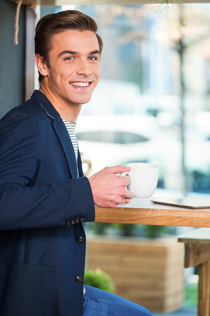 Bénéficiant d'un café frais. Beau jeune homme appréciant le café au café et regardant par-dessus l'épaule avec le sourire