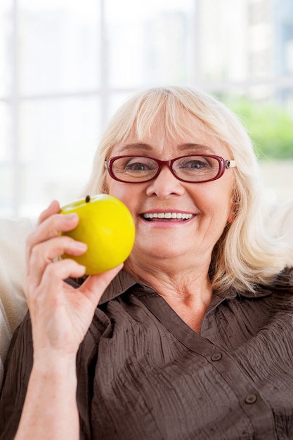 Bénéficiant d'un mode de vie sain. Cheerful senior woman holding apple et regardant la caméra alors qu'il était assis à la chaise