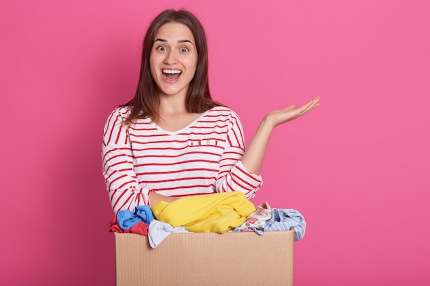 Bénévole Souriante Debout Près De La Boîte De Dons Contre Le Rose, Prenant Sa Main Sur Le Côté Et Garde La Bouche Ouverte