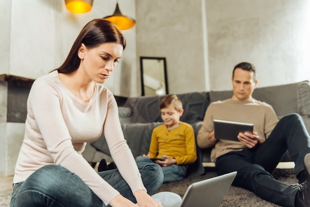 Besoin de se concentrer. Jolie jeune femme assise sur le sol et travaillant sur sa tablette, assise à distance de son fils et son mari à l'aide de leurs appareils