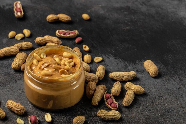 Beurre de cacahuète ou pâte dans un pot ouvert et cacahuètes dans la peau éparpillées sur un fond sombre bannière menu recette place pour texte vue de dessus