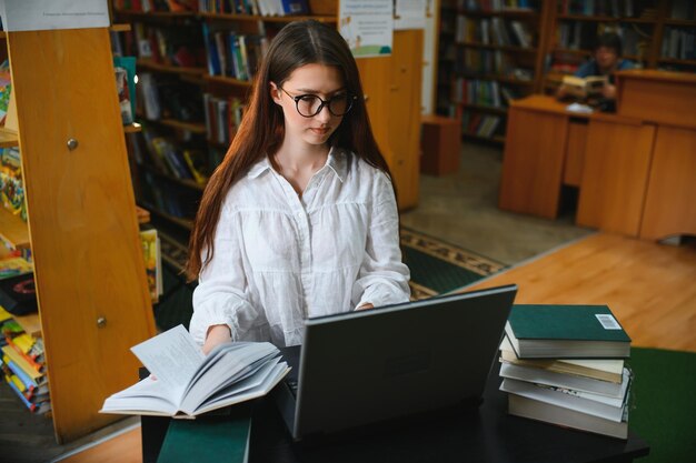 Bibliothèque universitaire Une belle fille caucasienne intelligente utilise un ordinateur portable Écrit des notes pour un essai sur papier Étude pour une affectation de classe Étudiants ciblés Apprentissage Étudier pour les examens universitaires