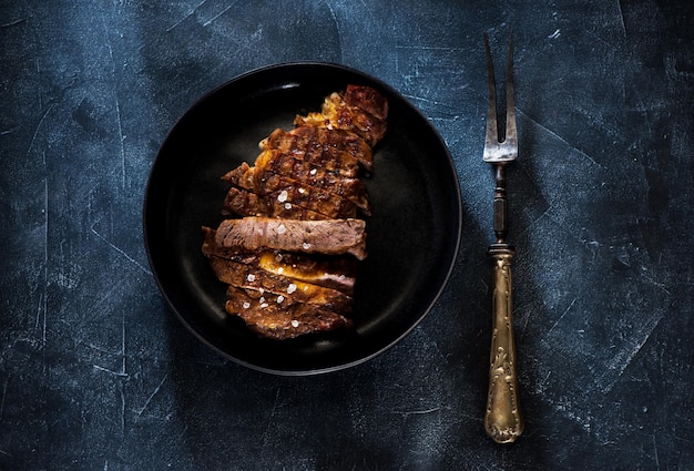 Bien cuit Steak Ribeye grillé dans une assiette et une fourchette antique sur un fond de béton foncé