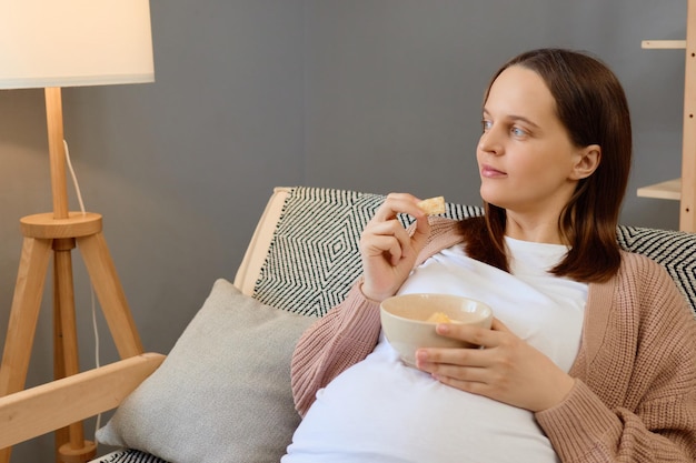 Bien-être grâce à la nutrition maternelle Femme enceinte calme et détendue assise sur le canapé tenant un bol de fruits frais mangeant une délicieuse collation regardant loin se reposant en profitant du temps à la maison
