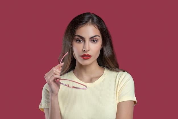 Bien-être. Sérieuse belle jeune femme aux cheveux longs en tshirt avec des lunettes à la main debout à l'intérieur