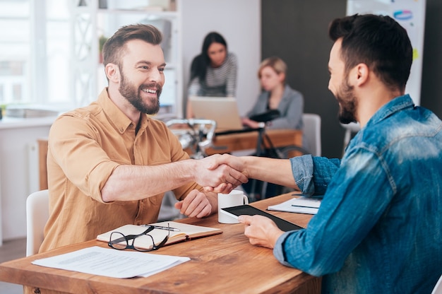 Bien fait! Deux jeunes hommes confiants se serrant la main et souriant alors qu'ils étaient assis au bureau avec deux personnes travaillant en arrière-plan