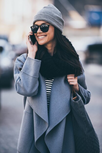 À bientôt! Belle jeune femme à lunettes de soleil parlant au téléphone portable et regardant loin avec le sourire tout en marchant à l'extérieur