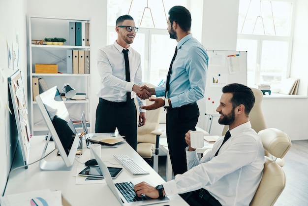 Bienvenue dans notre équipe ! Jeunes hommes modernes en tenues de soirée se serrant la main tout en travaillant au bureau