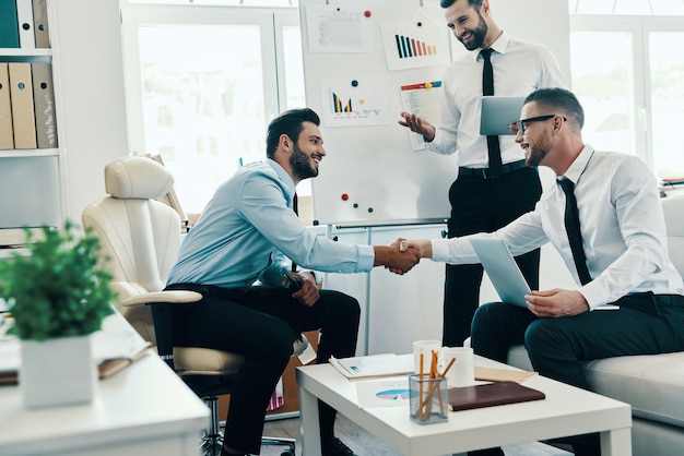 Bienvenue! Jeunes hommes modernes en tenues de soirée se serrant la main tout en travaillant au bureau