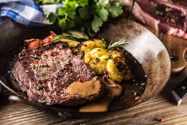 Bifteck de faux-filet de boeuf juteux pommes de terre rôties avec sauce légumes grillés sel poivre et herbes dans une poêle vintage.