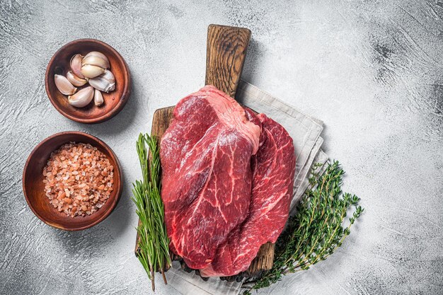 Biftecks de surlonge de viande de bœuf crue fraîche, herbes et épices autour d'une planche à découper. Fond blanc. Vue de dessus.