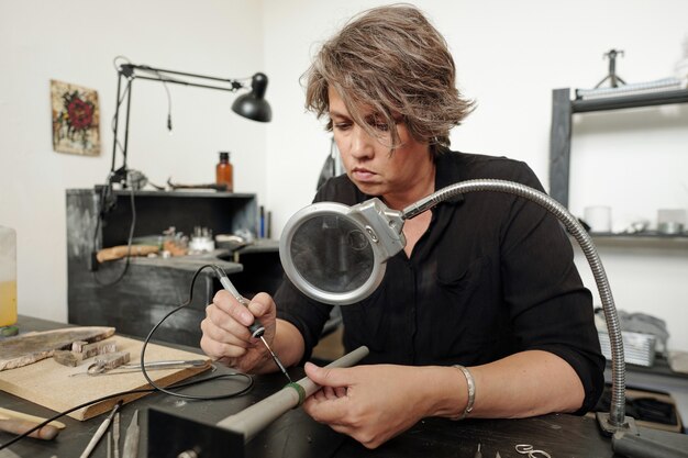 Bijoutier concentré assis dans un atelier avec des meubles noirs et des bijoux à souder...